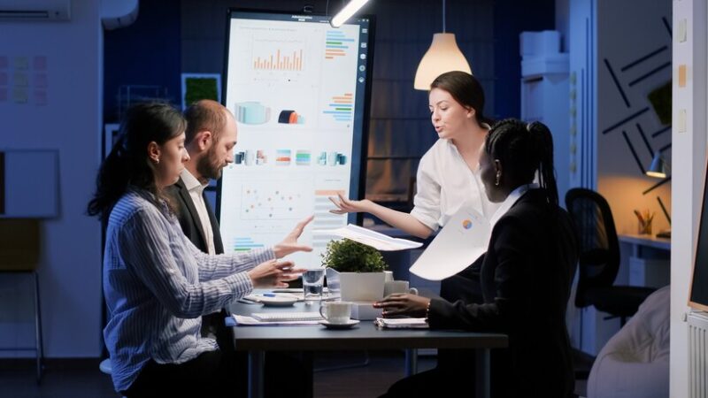 A diverse group of project managers and team members collaborating around a digital display of a PERT chart, engaged in discussion and planning, with laptops and tablets on the table, representing modern project management practices